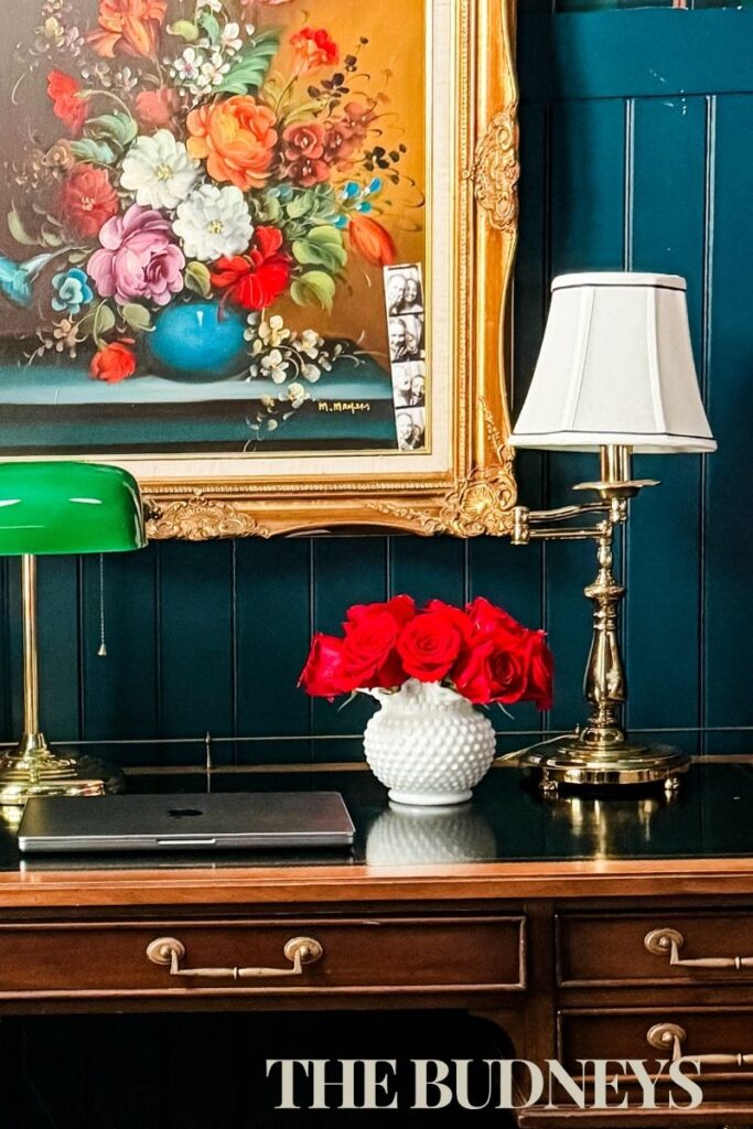 This image shows an antique feathertop wood desk. Styled with brass lamps, milk glass vase with red roses, and an antique floral oil painting.